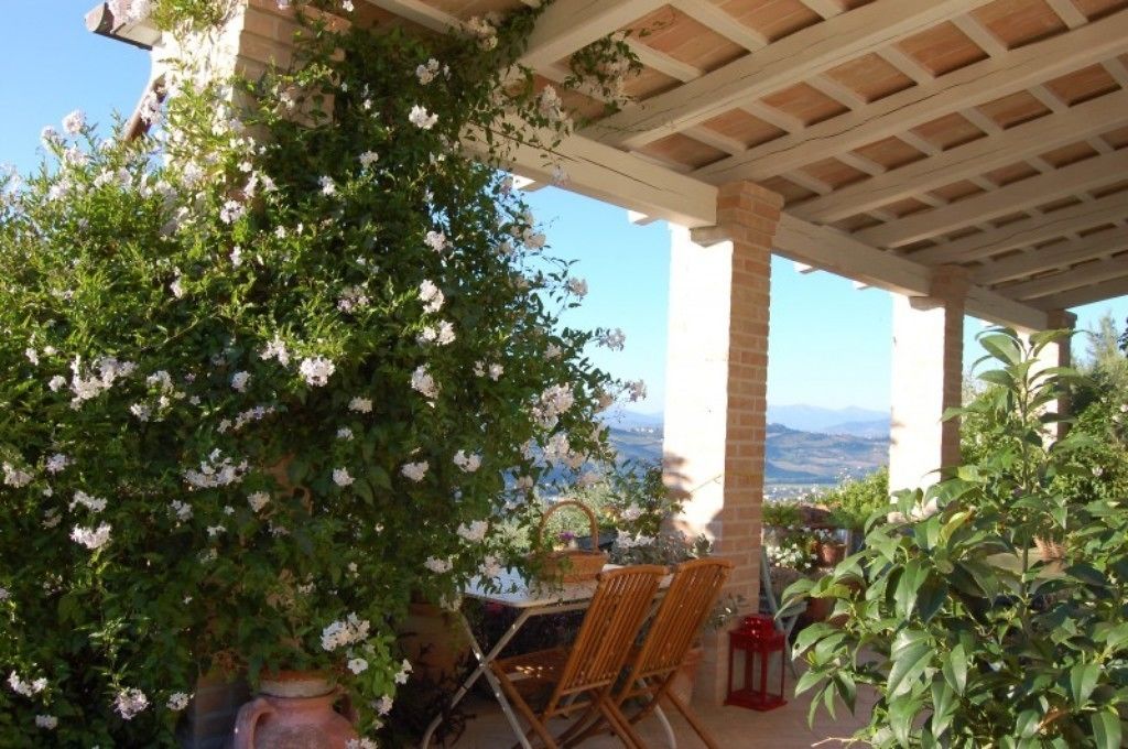 Un espace extérieur aménagé, abrité et doté de tables et de chaises, entouré d’une végétation luxuriante et de fleurs blanches.