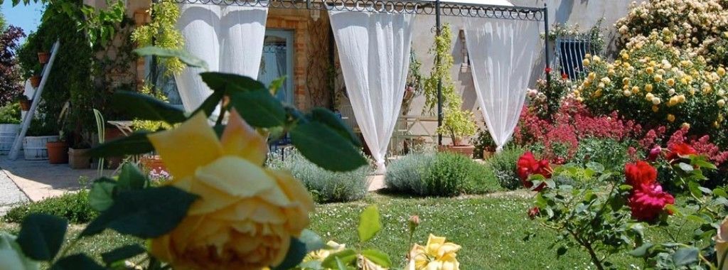 Un jardín con diversas flores y plantas, un edificio blanco con balcón y una valla.