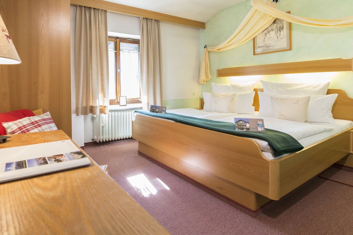 A hotel room with a wooden bed, a desk with a lamp, a window with curtains, and a headboard with curtains.