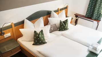 Uma cama de casal com lençóis brancos, duas almofadas estampadas em verde, uma cabeceira de madeira, uma mesa de cabeceira com abajur e um radiador.