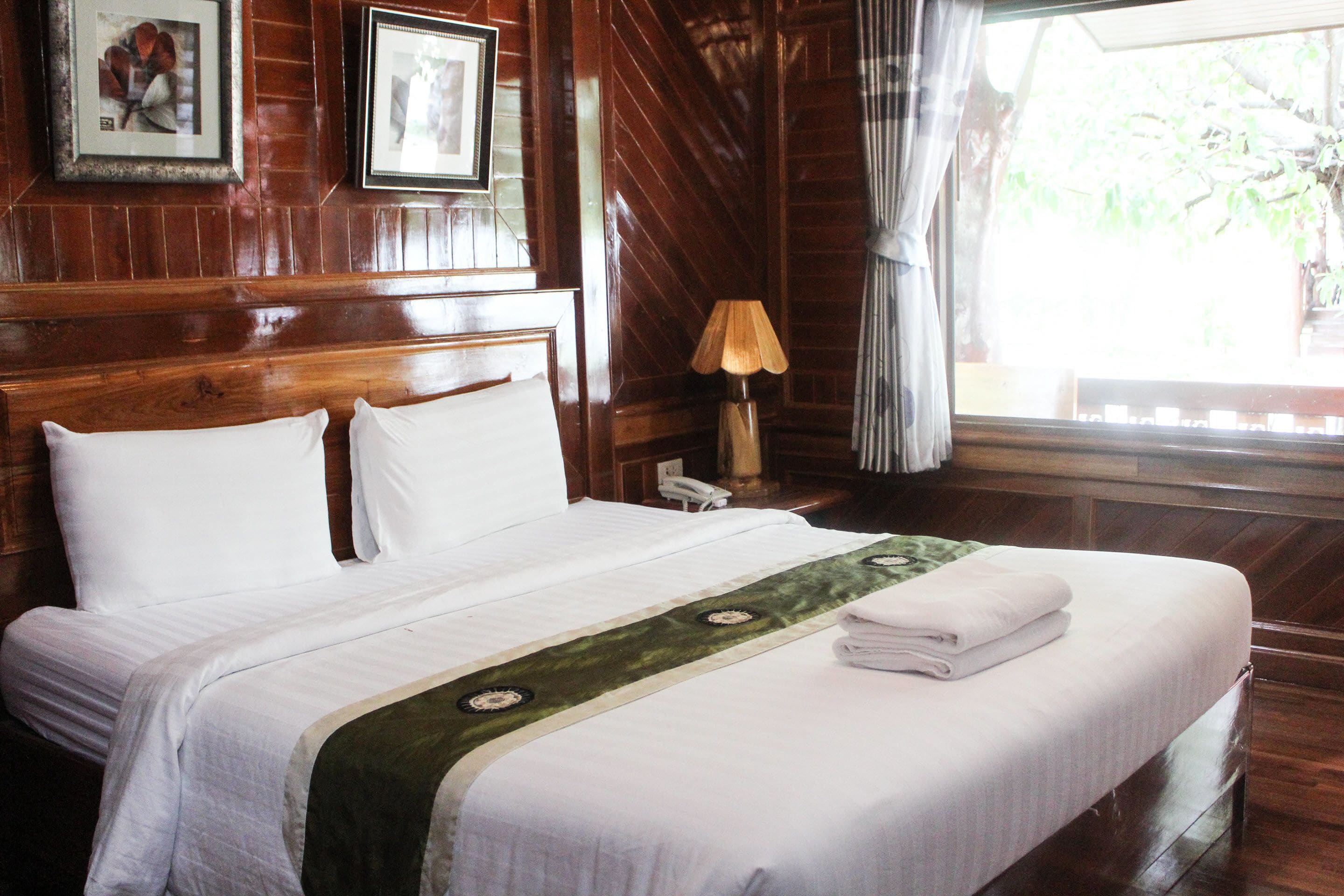 A neatly made bed with white linens, a green decorative runner, and a folded towel on the foot of the bed. There is a wooden headboard, a framed picture on the wall, and a lamp on a bedside table.