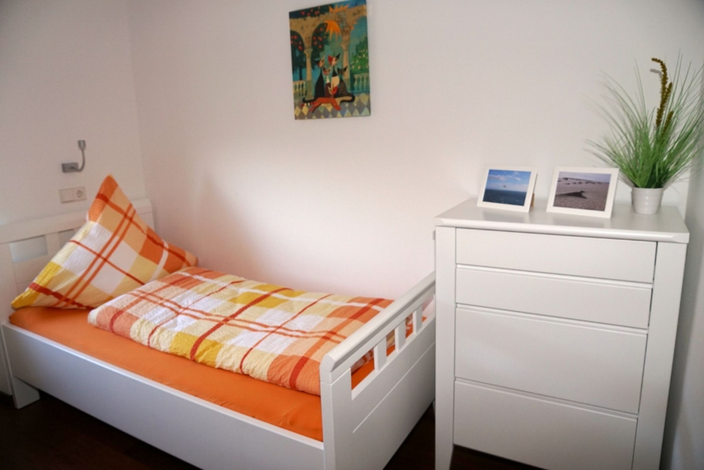 Een kamer met een enkel bed, een wit hoofdeinde, een oranje en witte geruit sprei, een witte nachtkastje met een plant in pot, en een aan de muur bevestigd fotolijstje.