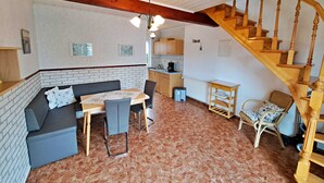 Un salon chaleureux avec un escalier en bois, un coin repas avec une table et des chaises, et une cuisine en arrière-plan.