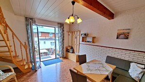 Un salon chaleureux avec un escalier en bois, un coin repas avec une table et des chaises, un canapé et une vue sur le balcon extérieur à travers une porte vitrée.