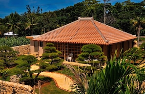 A traditional building with a tiled roof, surrounded by a landscaped garden with various trees and shrubs. - Kataakino Sato (Miyakojima)