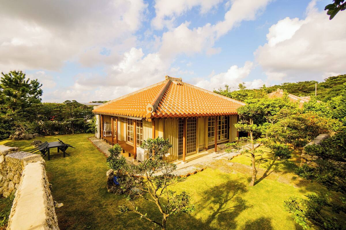 A traditional Japanese-style house with a tiled roof, surrounded by a well-maintained garden with a stone wall, a wooden deck, and a small table and chair set.