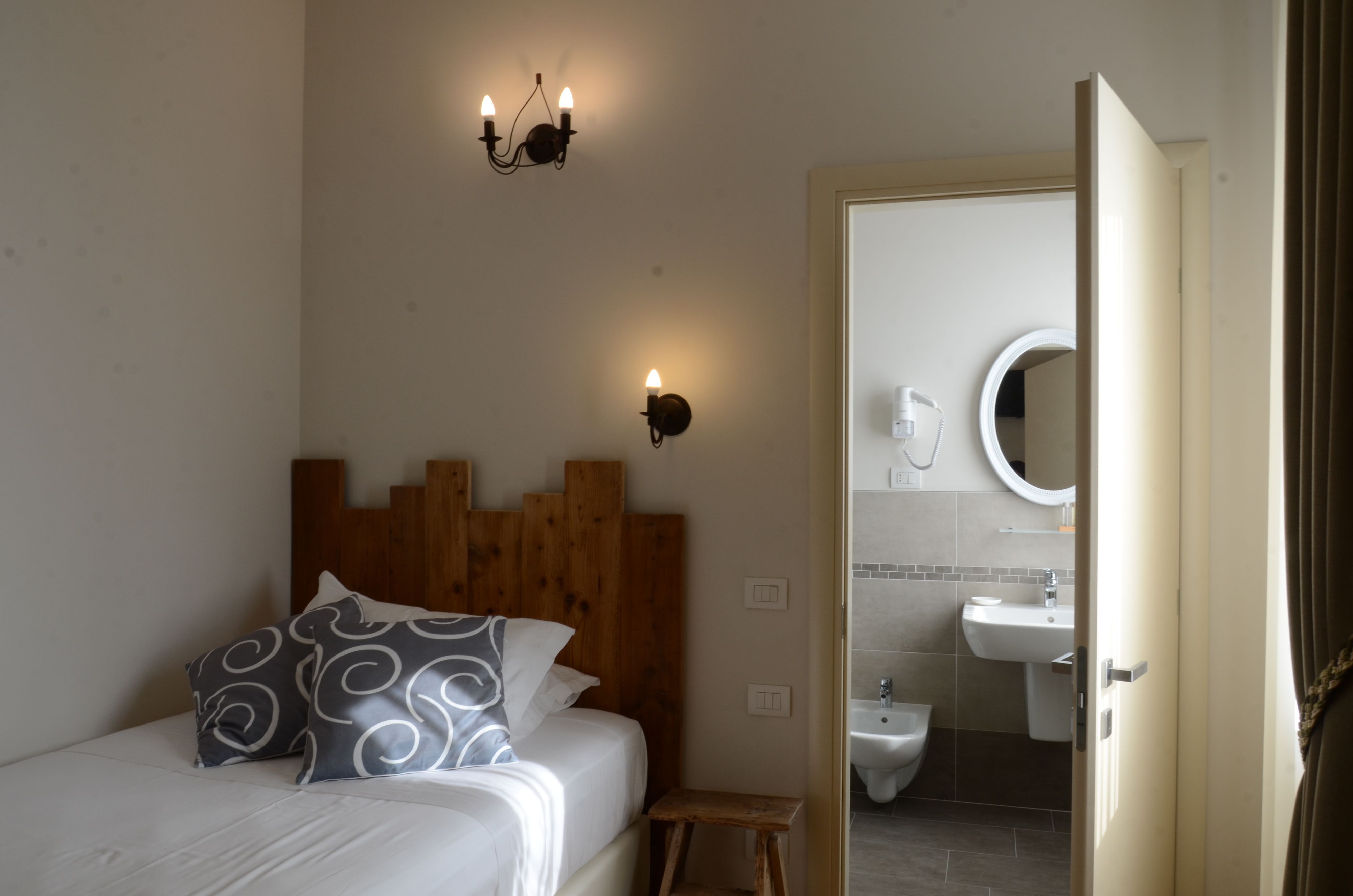 Une chambre à coucher avec une tête de lit en bois, un lit agrémenté d’oreillers à motifs, un petit tabouret en bois et une salle de bain visible à travers une porte ouverte.