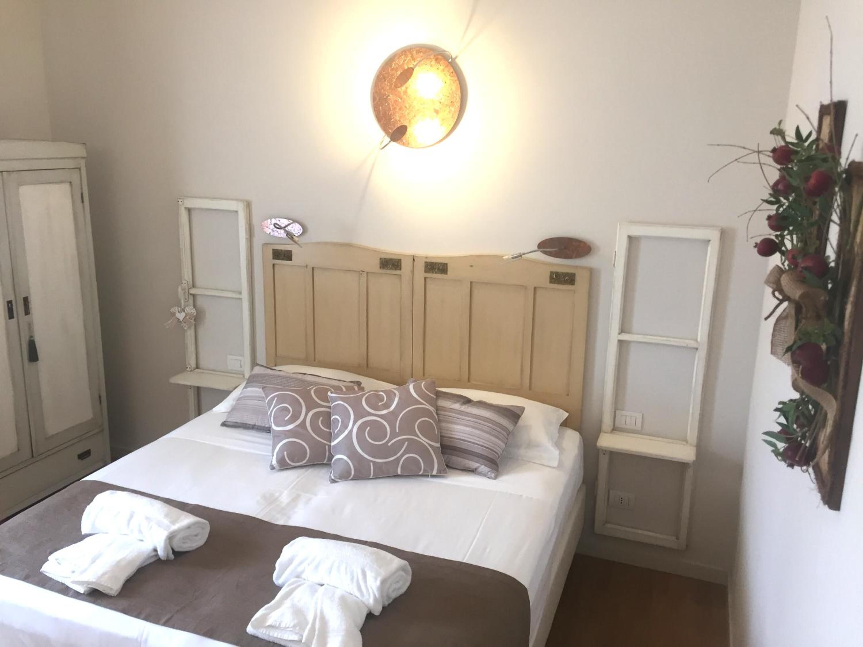 A neatly made bed with two pillows, a bedside table, a wall-mounted light fixture, and a decorative plant.