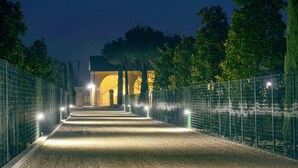Un chemin menant à un bâtiment dont l’entrée est illuminée d’une lumière chaleureuse, et qui est entouré d’une clôture en métal et d’arbres.