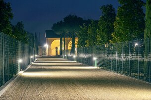 Un sentiero conduce a un edificio con un'entrata illuminata, circondato da un cancello in metallo e alberi.