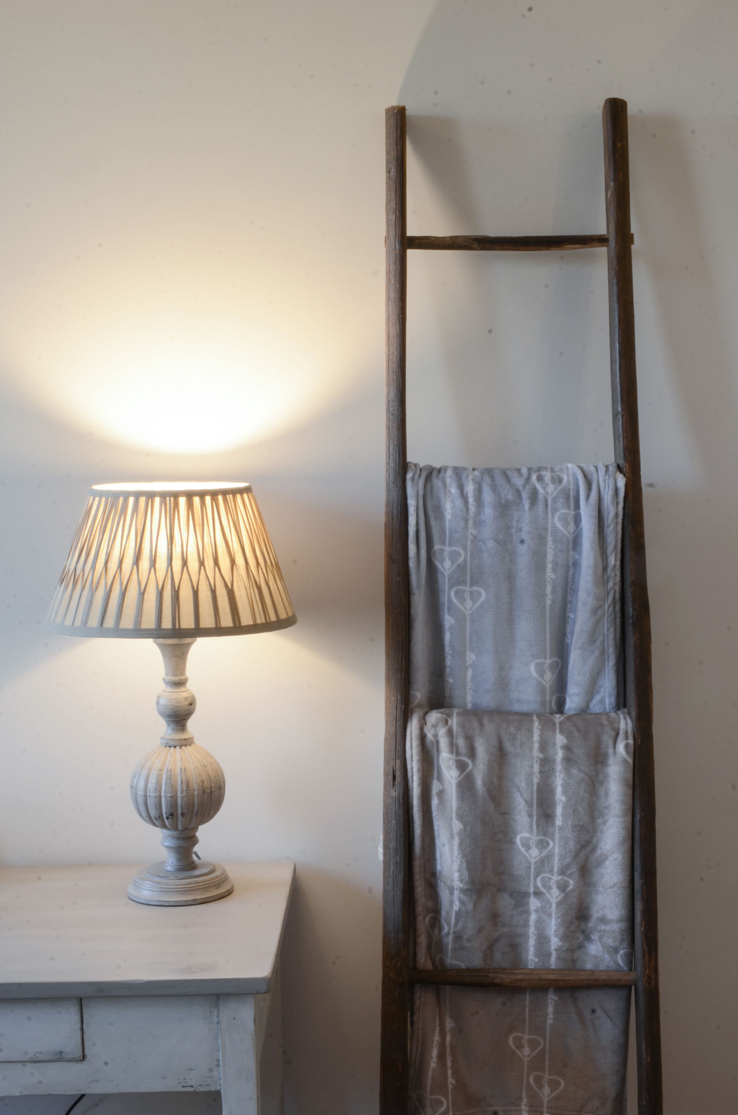A lamp with a ribbed shade on a white table, next to a wooden ladder leaning against a wall.