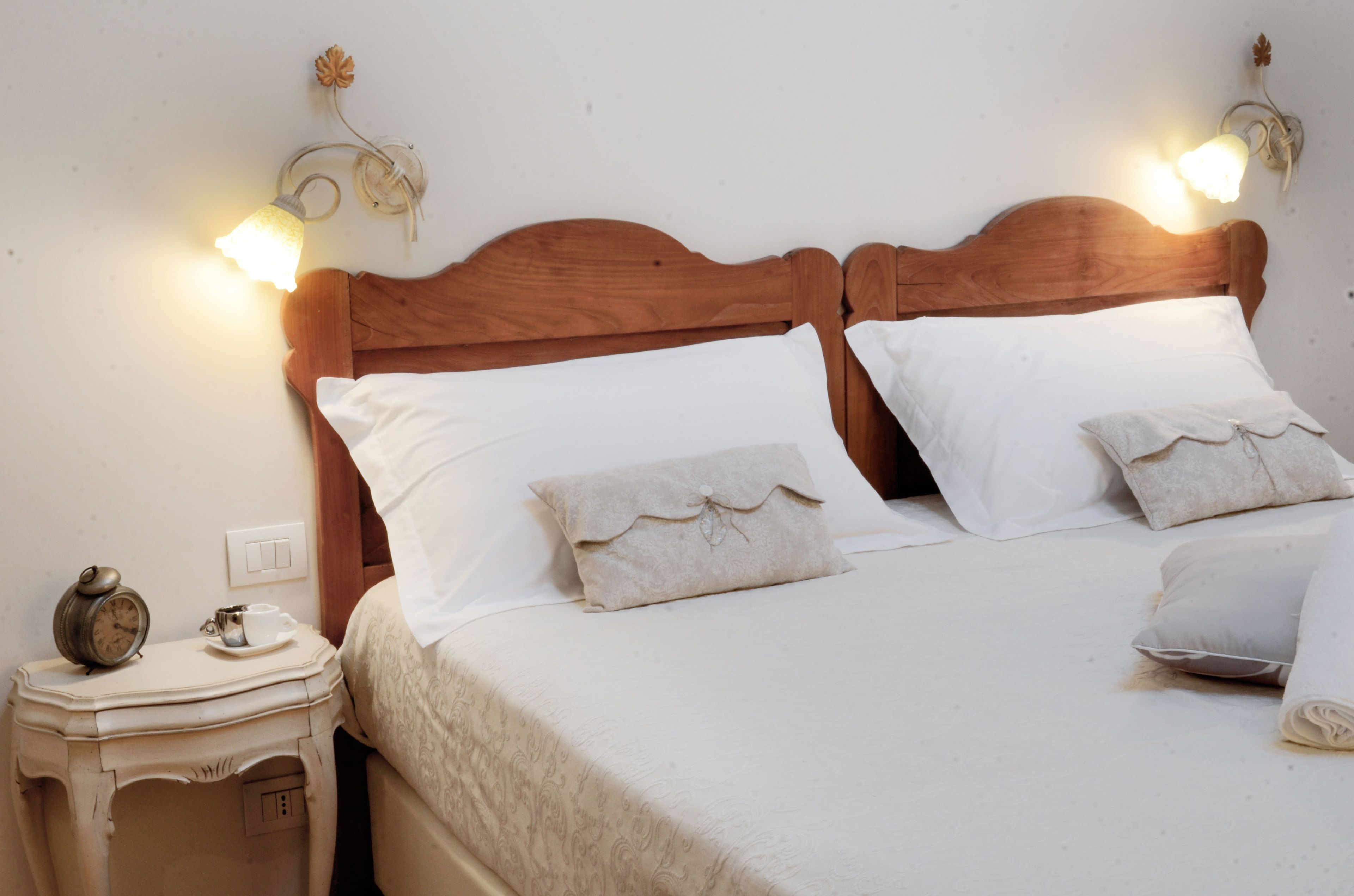 Een net opgemaakt bed met witte lakens en twee kussens, een nachtkastje met een lamp en een klein klokje, en wandlampjes.