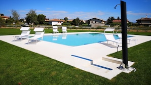 Une piscine entourée de chaises longues blanches et d’un poteau noir, avec une pelouse et des maisons en arrière-plan.