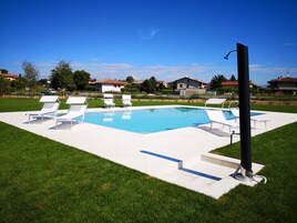 Una piscina con lettini bianchi e un palo nero, circondata da un'area verde e case sullo sfondo.