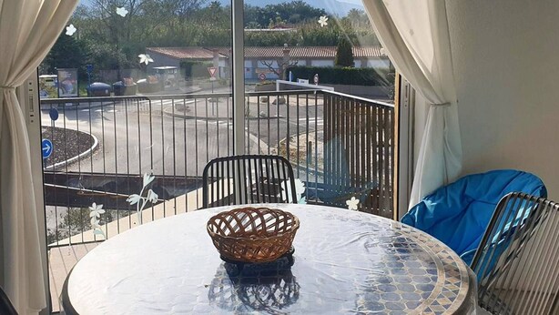 A balcony with a table and chairs, overlooking a street with a roundabout and mountains in the distance. - Cozy Studio with Balcony, Steps from the Beach - Sleeps 4, A/C, Parking (Argelès-sur-Mer)