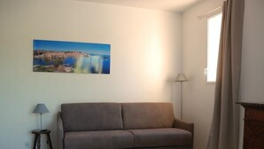 A living room with a sofa, a lamp, a small table with magazines, and a painting of a landscape on the wall. - Cozy Studio with Balcony, Steps from the Beach - Sleeps 4, A/C, Parking (Argelès-sur-Mer)