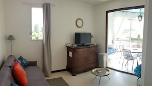 A living room with a sofa, a small coffee table, a TV on a wooden cabinet, and a view of an outdoor balcony with chairs. - Cozy Studio with Balcony, Steps from the Beach - Sleeps 4, A/C, Parking (Argelès-sur-Mer)