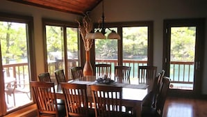 Un espace repas avec une table et des chaises en bois, de grandes fenêtres donnant sur des arbres et un plan d’eau, et un luminaire suspendu.