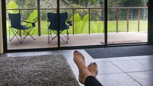 A view through a large window of a deck with two chairs, a grassy yard, and a forested area. - Tyenna River Cottage (Tyenna)