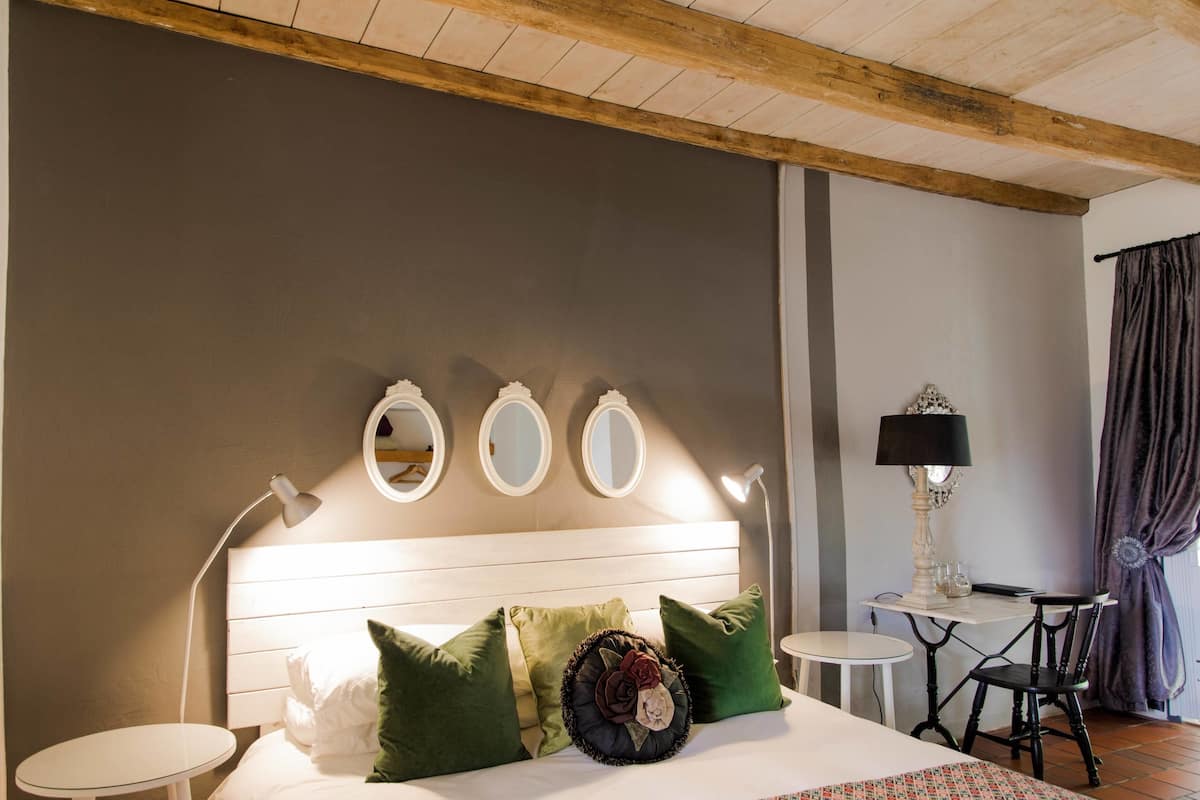 Un dormitorio con techo de madera, una cama con almohadas verdes y blancas, tres espejos redondos en la pared, una mesita de noche con una lámpara y una pequeña mesa con una silla.