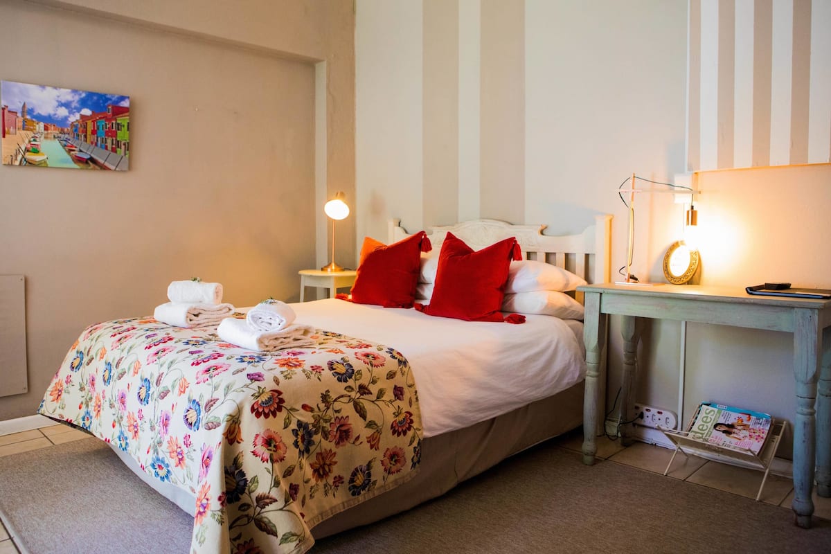 Una cama bien tendida con ropa de cama floral, una mesita de noche con una lámpara y un cuadro colgado en la pared.
