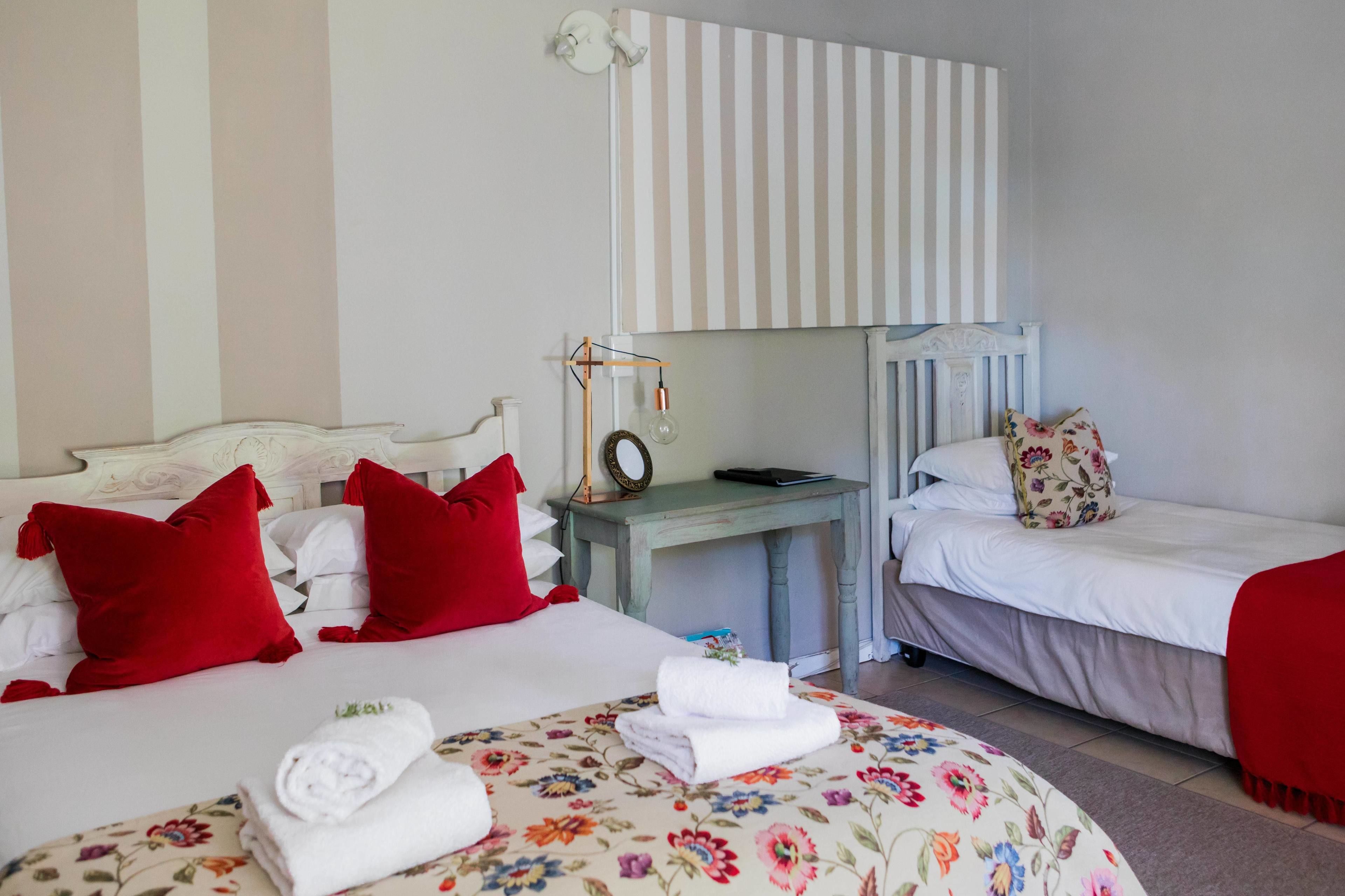 A hotel room with two beds, one with red pillows and the other with floral bedding. There is a small table with a lamp and a clock.