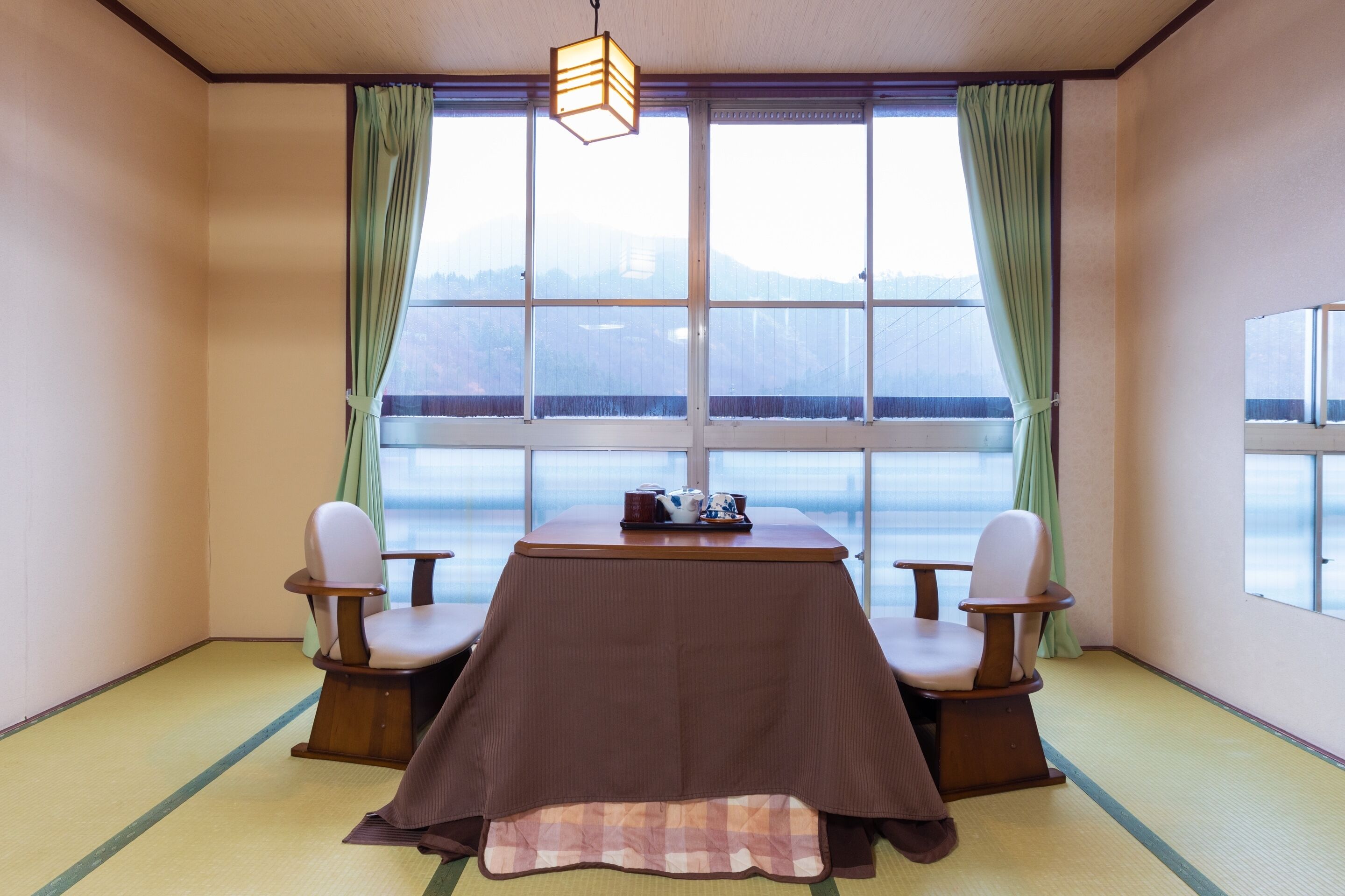 A room with tatami flooring, a low table with a brown tablecloth, two chairs, a hanging light fixture, and large windows with green curtains.