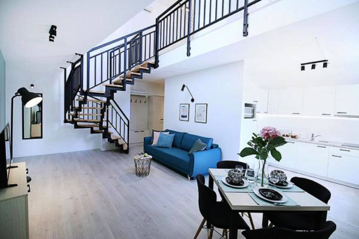 A modern living space with a staircase, a blue sofa, a dining table with chairs, and a kitchen area.