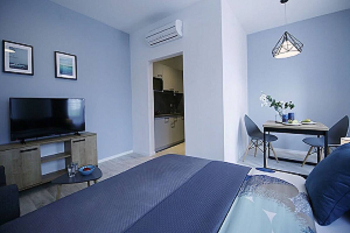 A modern hotel room with a large bed, a flat-screen TV, a dining area with a table and chairs, and a kitchenette in the background.