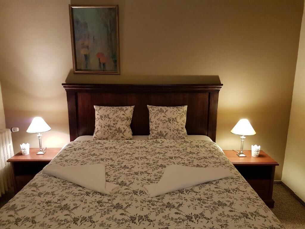 A hotel room with a bed, two bedside lamps, a painting on the wall, and a radiator.