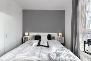 A modern bedroom with a large bed, two bedside tables with lamps, a fluffy rug, and a view of the city through sheer curtains. - Dream Holiday - Apartament Bastion Wałowa (Gdansk)