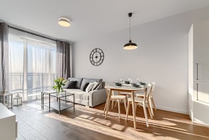 A modern living room with a sofa, coffee table, dining table, and chairs. There is a clock on the wall and a window with curtains.