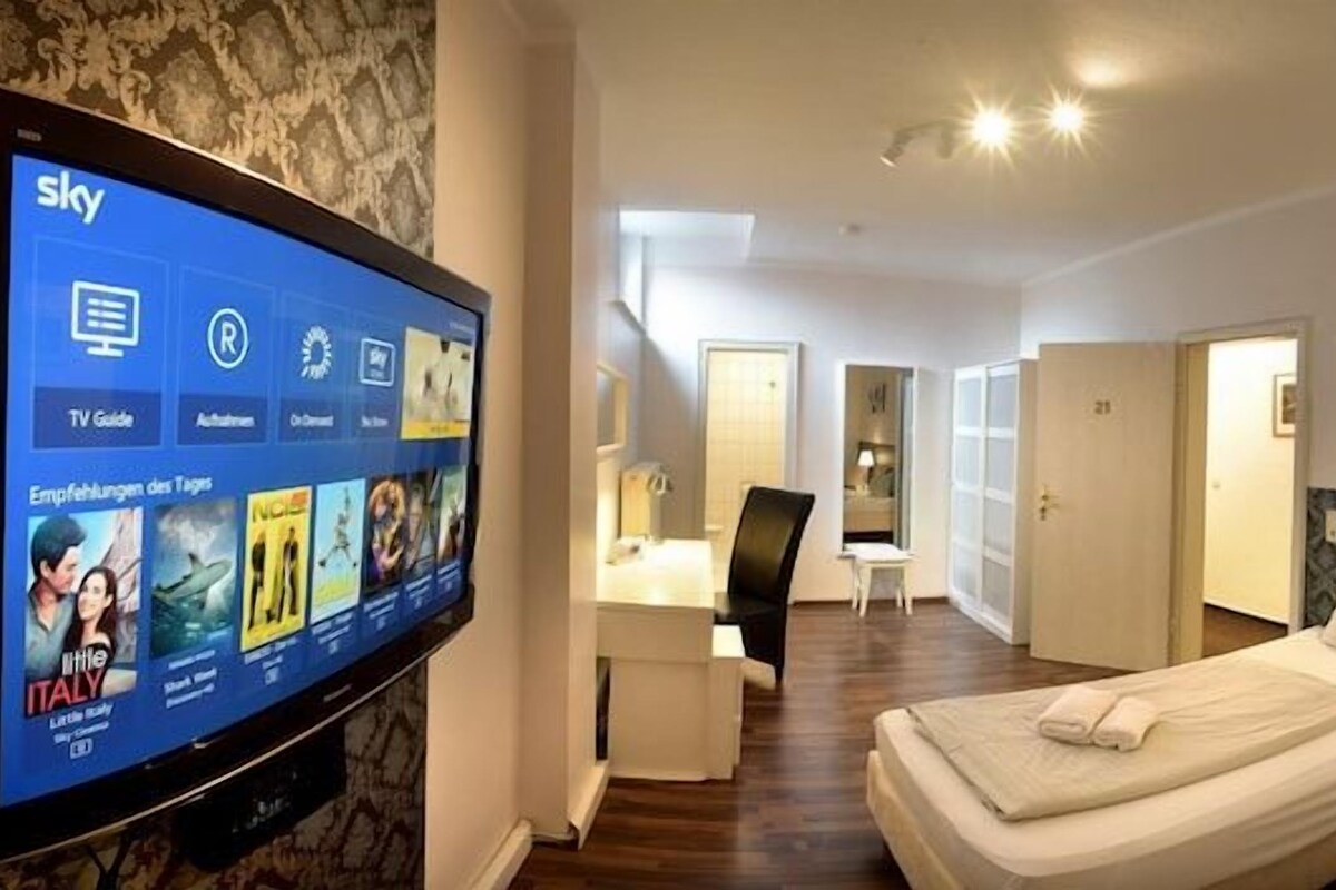 A hotel room with a TV displaying a TV guide, a bed with a headboard, a bedside table, and a wall-mounted lamp.