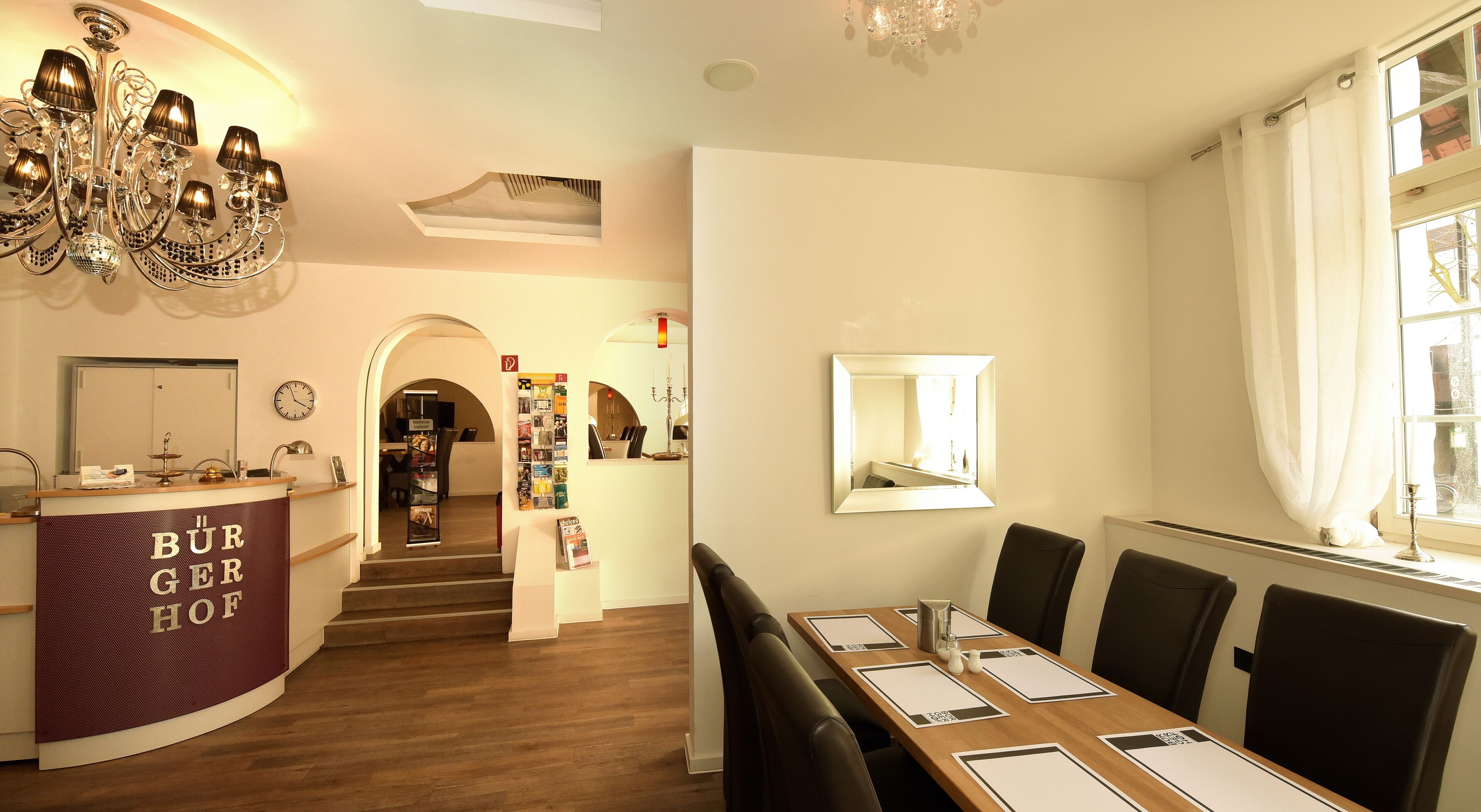 A dining area with a long table, chairs, and a chandelier. There is a reception desk with a sign that reads "Bürgerhof".