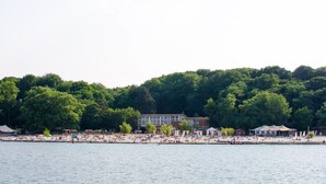 Una playa con gente tomando el sol y nadando, un edificio con un ventanal grande, y una colina boscosa al fondo.