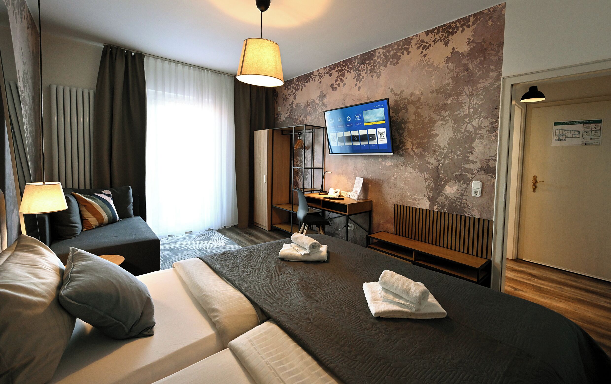 Ein Hotelzimmer mit Bett, Schreibtisch, Stuhl, Fernseher und einem Fenster mit Vorhängen.