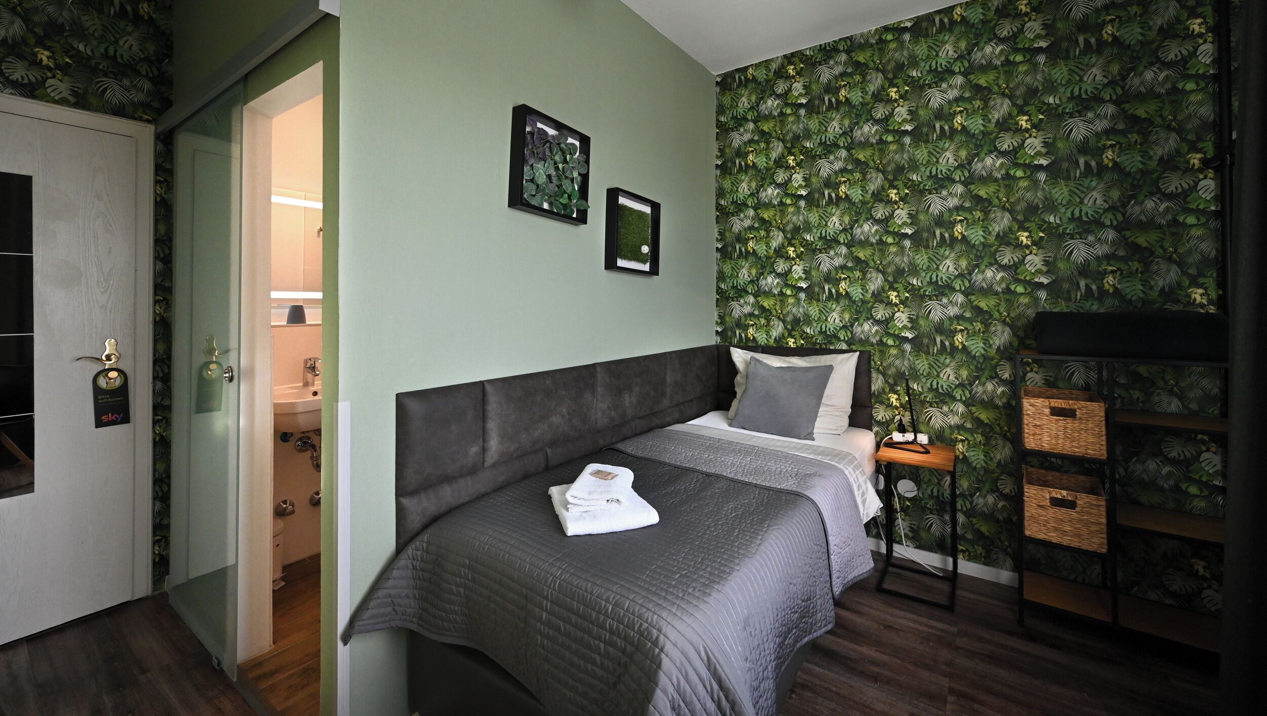A bedroom with a bed, a nightstand, a wall with a leafy pattern, and a door leading to a bathroom.