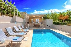 Un área de piscina con tumbonas, una mesa de madera y una piscina azul cristalina.