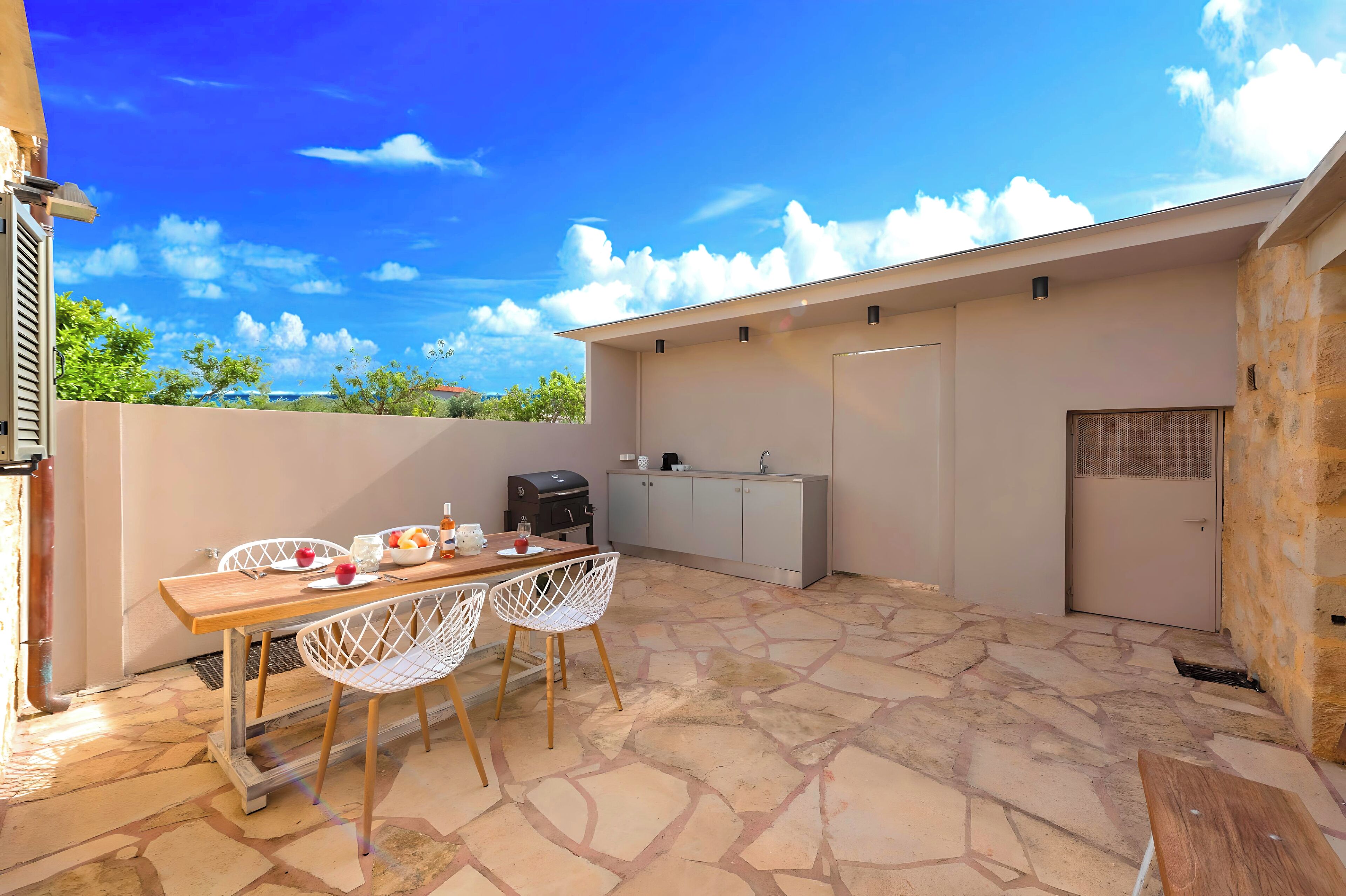 An outdoor patio with a stone floor, a wooden table with chairs, a grill, and a kitchen area with cabinets and a sink.