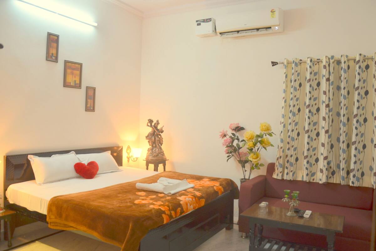 A hotel room with a bed, a sofa, a table, and an air conditioning unit.