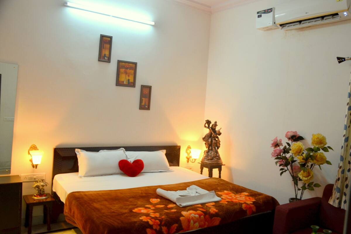 A hotel room with a bed, bedside lamps, a wall-mounted air conditioner, and a floral arrangement.