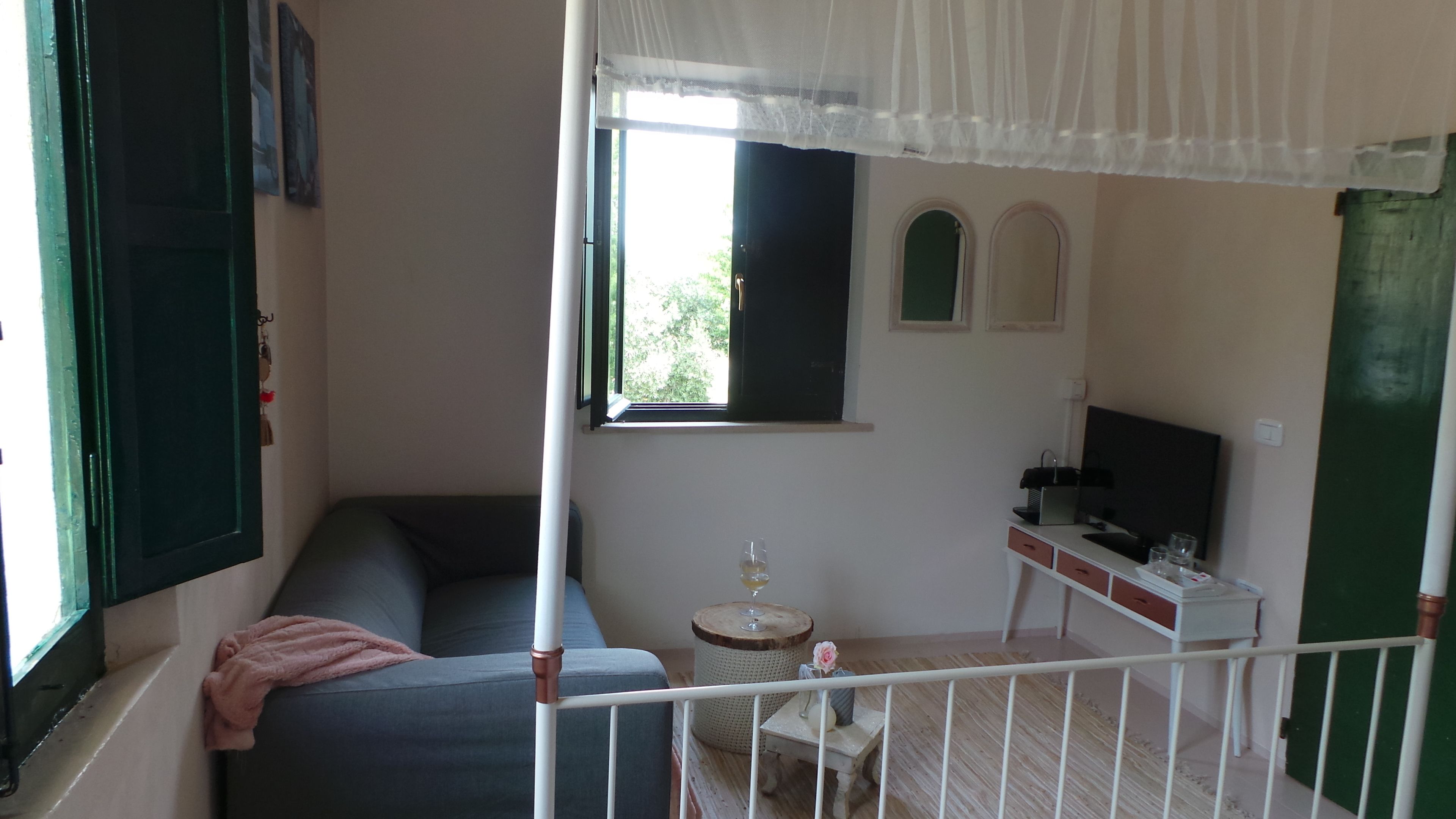 Een kamer met een bank, een klein rond tafeltje met een glas, een televisie op een standaard, en een raam met uitzicht op groen.