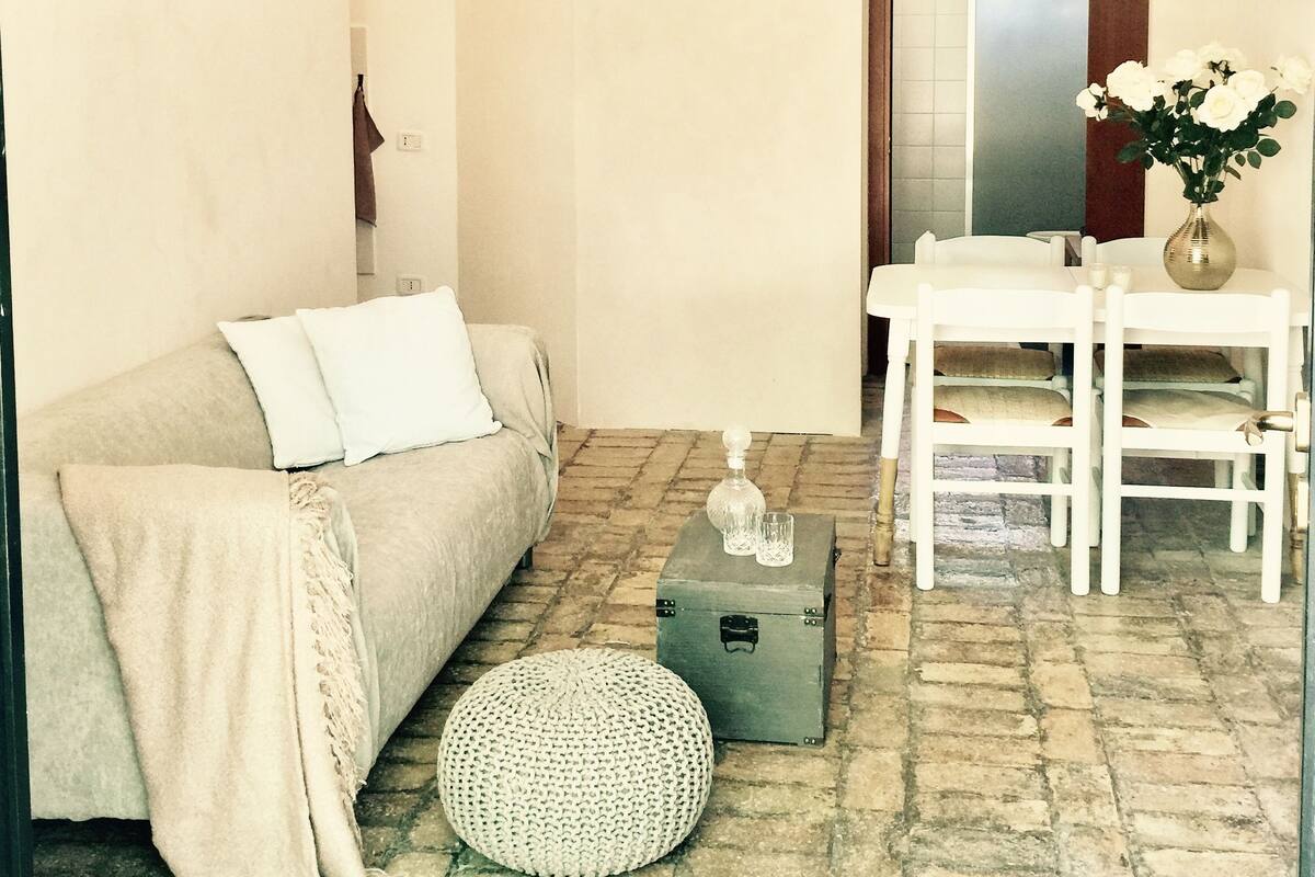 A cozy living room with a sofa, a small round ottoman, a dining table with chairs, a chandelier, and a wall clock.
