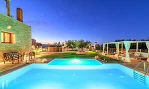 A swimming pool with a wooden deck, a pergola with white curtains, and a stone wall.