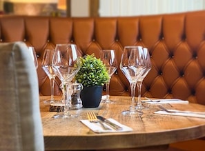 A dining table set with wine glasses, a small potted plant, and cutlery on a wooden table with a tufted leather bench in the background. - The Beeches (Ashby-de-la-Zouch)