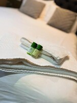 Una toalla blanca doblada con dos botellas verdes encima, sobre una cama con un almohadón blanco al fondo.