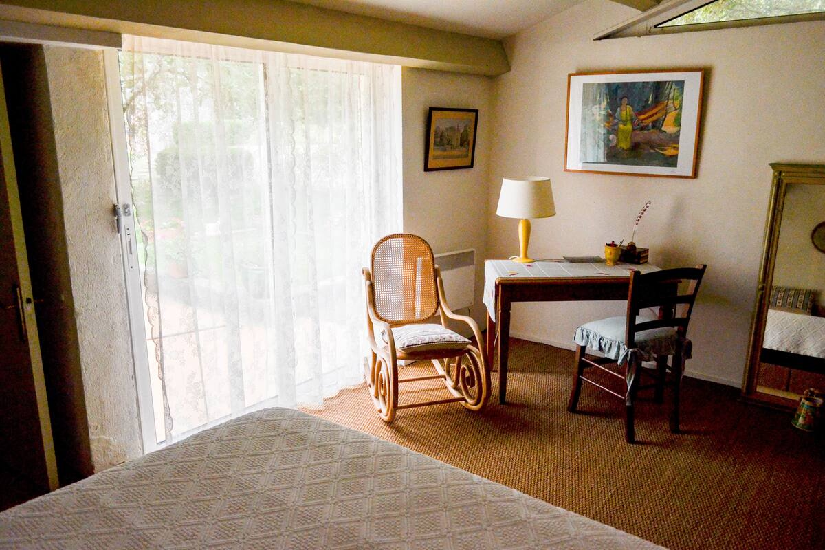 Une chambre d’hôtel avec un lit, une chaise en osier, un bureau avec une lampe et un tableau au mur.
