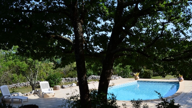 A pool area with a curved edge, surrounded by trees and potted plants. There are white lounge chairs and a small table. - Chambres d'hôtes Sous les Chênes (Lussan)