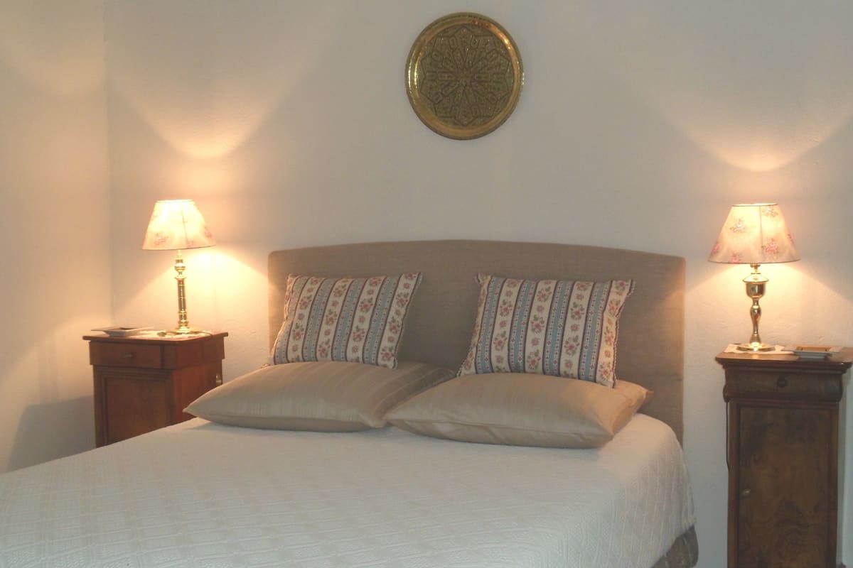 Un lit bien fait, agrémenté d’un coussin à motifs, deux tables de chevet avec des lampes et une assiette décorative accrochée au mur.