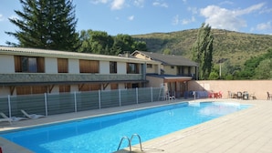 Une piscine avec une terrasse carrelée, des chaises longues et un bâtiment à l’arrière-plan, dont la façade est en pierre.